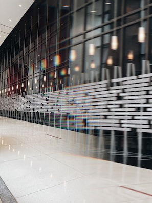 A modern interior space with a large reflective wall and an array of lights hanging from the ceiling. The wall features a series of horizontal patterns, and the reflection creates a distorted visual effect. The floor is polished and shiny, adding to the sleek design.