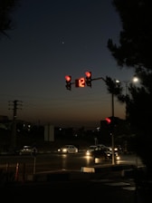 A night scene featuring a traffic signal displaying a red light with the number 12 on a digital timer. Cars are visible on the road, some with headlights on. The sky is dark, transitioning to a dim twilight at the horizon. Silhouetted trees and power lines are present in the background.