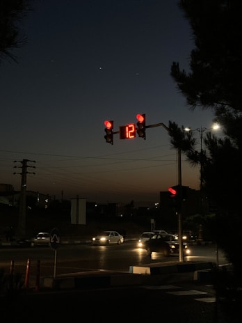 A night scene featuring a traffic signal displaying a red light with the number 12 on a digital timer. Cars are visible on the road, some with headlights on. The sky is dark, transitioning to a dim twilight at the horizon. Silhouetted trees and power lines are present in the background.