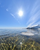 A bright sun shines in a clear blue sky over a tranquil body of water. The water is clear, revealing a rocky bottom with colorful reflections. Large chunks of ice are visible on the right side, suggesting a cold environment.