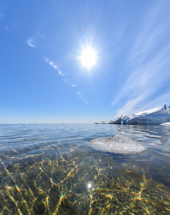 A bright sun shines in a clear blue sky over a tranquil body of water. The water is clear, revealing a rocky bottom with colorful reflections. Large chunks of ice are visible on the right side, suggesting a cold environment.