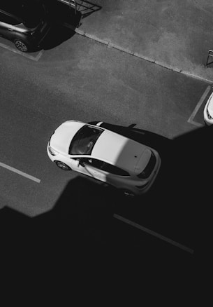 Aerial view of a white car driving on a dark road with distinct shadows cast by nearby buildings. The car appears to be in motion, with clear lines marking the lanes on the road. A parked car is visible nearby, and the image is rendered in black and white, accentuating the contrast between light and shadow.