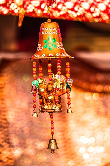 A colorful hanging decoration featuring a bell-shaped top with intricate patterns in red and green. Below are tiny bells and beads in red and gold, with small ornamental elephants and vibrant accents. The background is softly blurred with warm bokeh lights.