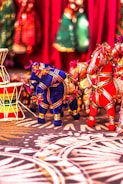 Colorful fabric horse figurines are arranged on a patterned surface. They are adorned with intricate thread work in various colors. In the background, there is a red fabric backdrop with blurred shapes that appear to be traditional clothing or decorative items. A colorful drum-like object is placed on the left side.