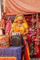 A person dressed in traditional colorful attire is standing in front of a vibrantly decorated background. They are interacting with an electronic device that resembles an amplifier or speaker. The setting includes ornate fabrics with red and gold patterns and various decorative items that suggest a cultural or festival environment.