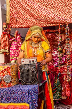 A person dressed in traditional colorful attire is standing in front of a vibrantly decorated background. They are interacting with an electronic device that resembles an amplifier or speaker. The setting includes ornate fabrics with red and gold patterns and various decorative items that suggest a cultural or festival environment.