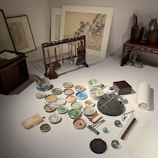 A set of traditional painting tools arranged neatly on a table, including a variety of brushes hanging on a stand, multiple small bowls containing different colored pigments, and drawing papers. In the background, framed artworks featuring sketches and paintings of landscapes. The scene conveys a sense of artistic creation in a serene workspace.