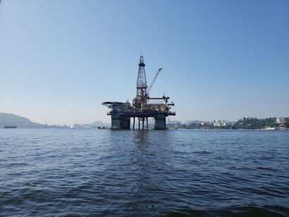 An offshore oil rig stands in the middle of a body of water, with a clear blue sky in the background. The structure is supported by multiple pillars and features a derrick and cranes, suggesting it is actively used for oil extraction. Surrounding the rig, there are distant cityscapes and verdant hills, indicating its proximity to a coastal area.