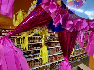 Brightly colored piñatas with shiny pink wrapping and yellow tassels are hanging indoors against the backdrop of a grocery store. Numerous shelves are filled with assorted products including jars and cans organized neatly.