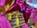 Brightly colored piñatas with shiny pink wrapping and yellow tassels are hanging indoors against the backdrop of a grocery store. Numerous shelves are filled with assorted products including jars and cans organized neatly.