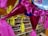 Brightly colored piñatas with shiny pink wrapping and yellow tassels are hanging indoors against the backdrop of a grocery store. Numerous shelves are filled with assorted products including jars and cans organized neatly.