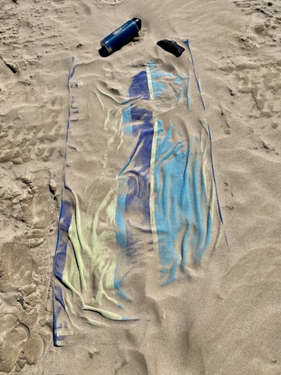 A sandy beach with an unfolded towel, partially covered by sand, displaying blue and green patterns. A blue water bottle and a pair of sunglasses are placed on the sand nearby.
