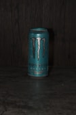 A can of Monster Energy drink, specifically the 'Ultra Fiesta Mango' flavor, is placed on a dark surface. The can is primarily blue with the iconic Monster logo in black and silver. The background appears to be a textured dark material, possibly wood or stone.