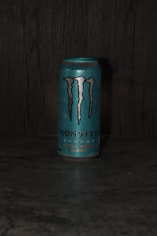 A can of Monster Energy drink, specifically the 'Ultra Fiesta Mango' flavor, is placed on a dark surface. The can is primarily blue with the iconic Monster logo in black and silver. The background appears to be a textured dark material, possibly wood or stone.