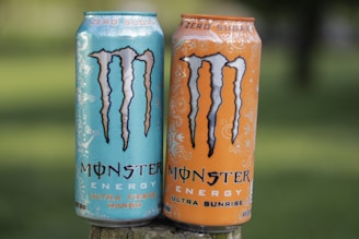 Two cans of Monster Energy drink stand side by side on a wooden surface against a blurred natural background. The can on the left is blue with 'Ultra Fiesta Mango' written on it, while the can on the right is orange with 'Ultra Sunrise' written on it. Both cans are labeled 'Zero Sugar' and have a stylized 'M' logo prominently displayed.