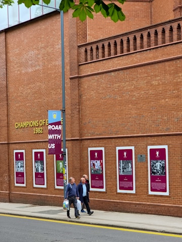 A brick building displays numerous framed images and text related to a sports team or event, indicated by the words 'Champions of Europe 1982'. Several people walk along the sidewalk beside the building, and a lamp post with a banner saying 'Roar With Us' is partially visible. Green leaves from a tree overhang the scene in the top part of the image.