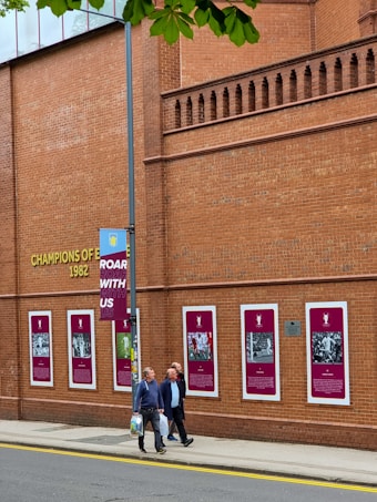 A brick building displays numerous framed images and text related to a sports team or event, indicated by the words 'Champions of Europe 1982'. Several people walk along the sidewalk beside the building, and a lamp post with a banner saying 'Roar With Us' is partially visible. Green leaves from a tree overhang the scene in the top part of the image.