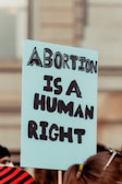 A sign being held at a protest that reads 'Abortion is a Human Right' in bold, black letters on a light blue background. The sign is in focus, while the background appears to be a blurred crowd and a building.