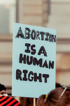 A sign being held at a protest that reads 'Abortion is a Human Right' in bold, black letters on a light blue background. The sign is in focus, while the background appears to be a blurred crowd and a building.