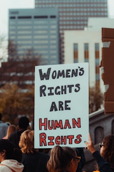A group of people participating in a protest or rally, with a prominent sign in the foreground that reads 'Women's Rights Are Human Rights'. The background features tall buildings, suggesting an urban setting.