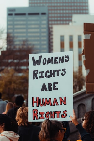 A group of people participating in a protest or rally, with a prominent sign in the foreground that reads 'Women's Rights Are Human Rights'. The background features tall buildings, suggesting an urban setting.