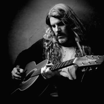 A black and white portrait of a person with long hair playing an acoustic guitar. The individual appears focused and deeply engaged with the music. The lighting casts dramatic shadows, highlighting facial features and creating a moody atmosphere.