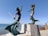 Two bronze mermaid sculptures are mounted on elevated concrete pedestals by the seaside. The male mermaid holds a trident and is posed dynamically, reaching his arm behind him. The female mermaid is positioned to his right, with one hand placed on her head and the other extended as if gesturing. The sea and a clear blue sky are visible in the background, with some distant buildings and a slight mountain range along the coastline.