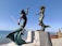 Two bronze mermaid sculptures are mounted on elevated concrete pedestals by the seaside. The male mermaid holds a trident and is posed dynamically, reaching his arm behind him. The female mermaid is positioned to his right, with one hand placed on her head and the other extended as if gesturing. The sea and a clear blue sky are visible in the background, with some distant buildings and a slight mountain range along the coastline.
