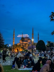 A vibrant evening scene in a park with people gathered, sitting on the grass and benches. In the background, an illuminated mosque stands with minarets, and a large text display reads 'MUHAMMED RESULULLAH'. Trees and lampposts are scattered throughout the area.