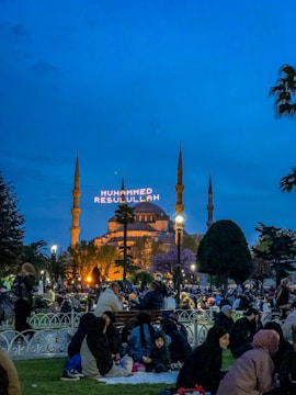A vibrant evening scene in a park with people gathered, sitting on the grass and benches. In the background, an illuminated mosque stands with minarets, and a large text display reads 'MUHAMMED RESULULLAH'. Trees and lampposts are scattered throughout the area.