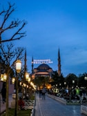A wide walkway lined with ornate street lamps leads towards a large, historic mosque with multiple minarets under a dusk sky. The mosque is lit up, and an illuminated message in Arabic script appears between the minarets. People are casually strolling or sitting in the park-like area surrounding the mosque.