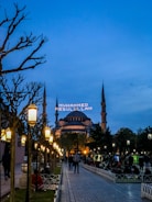 A wide walkway lined with ornate street lamps leads towards a large, historic mosque with multiple minarets under a dusk sky. The mosque is lit up, and an illuminated message in Arabic script appears between the minarets. People are casually strolling or sitting in the park-like area surrounding the mosque.