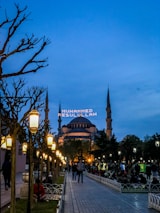 A wide walkway lined with ornate street lamps leads towards a large, historic mosque with multiple minarets under a dusk sky. The mosque is lit up, and an illuminated message in Arabic script appears between the minarets. People are casually strolling or sitting in the park-like area surrounding the mosque.