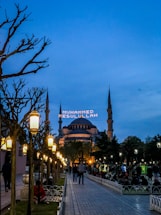 A wide walkway lined with ornate street lamps leads towards a large, historic mosque with multiple minarets under a dusk sky. The mosque is lit up, and an illuminated message in Arabic script appears between the minarets. People are casually strolling or sitting in the park-like area surrounding the mosque.