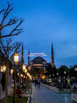 A wide walkway lined with ornate street lamps leads towards a large, historic mosque with multiple minarets under a dusk sky. The mosque is lit up, and an illuminated message in Arabic script appears between the minarets. People are casually strolling or sitting in the park-like area surrounding the mosque.