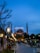 A wide walkway lined with ornate street lamps leads towards a large, historic mosque with multiple minarets under a dusk sky. The mosque is lit up, and an illuminated message in Arabic script appears between the minarets. People are casually strolling or sitting in the park-like area surrounding the mosque.