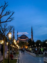 A wide walkway lined with ornate street lamps leads towards a large, historic mosque with multiple minarets under a dusk sky. The mosque is lit up, and an illuminated message in Arabic script appears between the minarets. People are casually strolling or sitting in the park-like area surrounding the mosque.