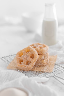 A stack of sugar-coated pastries rests on a piece of parchment paper atop a metal cooling rack. In the background, a glass bottle of milk is slightly out of focus, accompanied by a soft, white cloth.