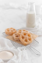 A few sugar-coated pastries with a lattice shape are placed on a metal cooling rack. The background features a glass bottle filled with milk. A white cloth and a small bowl of sugar are also visible, creating a soft, inviting baking scene.