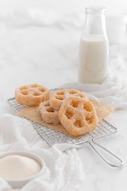 A few sugar-coated pastries with a lattice shape are placed on a metal cooling rack. The background features a glass bottle filled with milk. A white cloth and a small bowl of sugar are also visible, creating a soft, inviting baking scene.