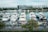 Several luxury yachts are docked in a marina, with a large number of smaller boats neatly arranged in front. Modern buildings and lush green trees line the background, and the water is calm, reflecting the sky and structures.