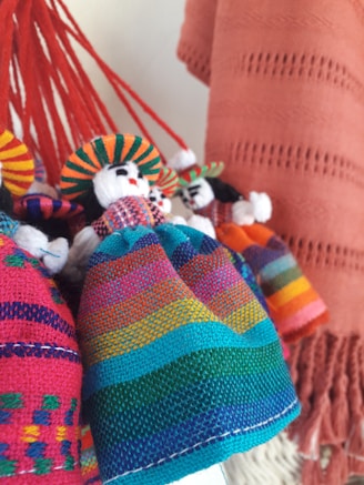A collection of handmade dolls adorned in colorful, traditional woven garments, featuring vibrant patterns. The dolls have bright hats and distinct facial features, with backgrounds of similar textile material.