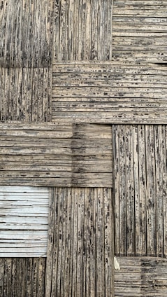 A textured surface consisting of interwoven bamboo strips arranged in a square pattern. The bamboo appears weathered, with a mix of dark and light brown shades, and some areas show signs of aging or wear.