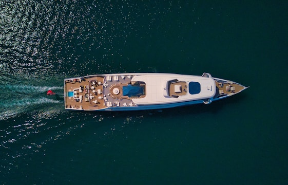 Aerial view of a luxurious yacht on a calm sea, featuring an expansive deck with seating areas and a central pool. The wake of the vessel is visible behind it, indicating movement. The water is a deep blue-green color, contrasting with the yacht's white hull.