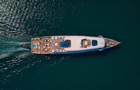 Aerial view of a luxurious yacht on a calm sea, featuring an expansive deck with seating areas and a central pool. The wake of the vessel is visible behind it, indicating movement. The water is a deep blue-green color, contrasting with the yacht's white hull.