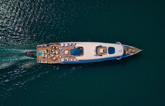 Aerial view of a luxurious yacht on a calm sea, featuring an expansive deck with seating areas and a central pool. The wake of the vessel is visible behind it, indicating movement. The water is a deep blue-green color, contrasting with the yacht's white hull.