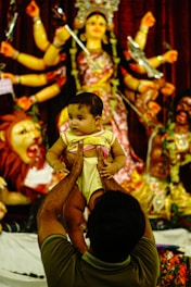 A person is holding a baby up in front of a colorful and elaborate statue of a multi-armed deity, possibly representing a Hindu goddess. The scene suggests a cultural or religious setting.