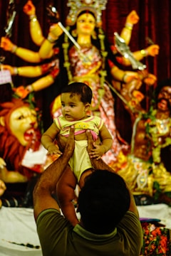 A person is holding a baby up in front of a colorful and elaborate statue of a multi-armed deity, possibly representing a Hindu goddess. The scene suggests a cultural or religious setting.