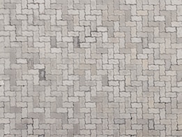 An arrangement of light gray interlocking paving stones forms a repetitive pattern. The pavers are rectangular with irregular, wavy edges, creating a uniform and structured surface.