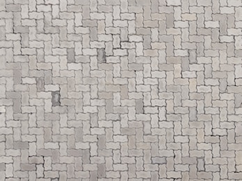 An arrangement of light gray interlocking paving stones forms a repetitive pattern. The pavers are rectangular with irregular, wavy edges, creating a uniform and structured surface.